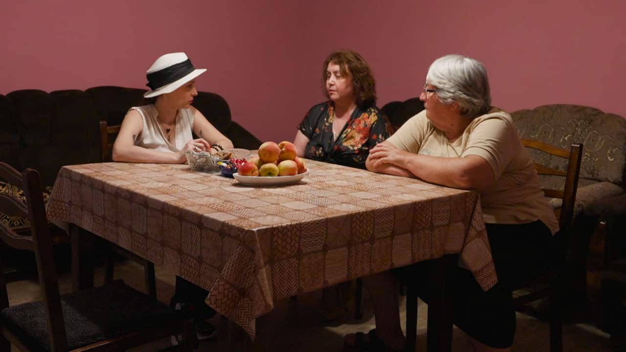 Three women sit at a table with a plate of apples and lollies, having a conversation. The woman on the left is wearing a white hat with a black ribbon, a middle-aged woman is in the middle, and an elderly woman with gray hair is on the right.