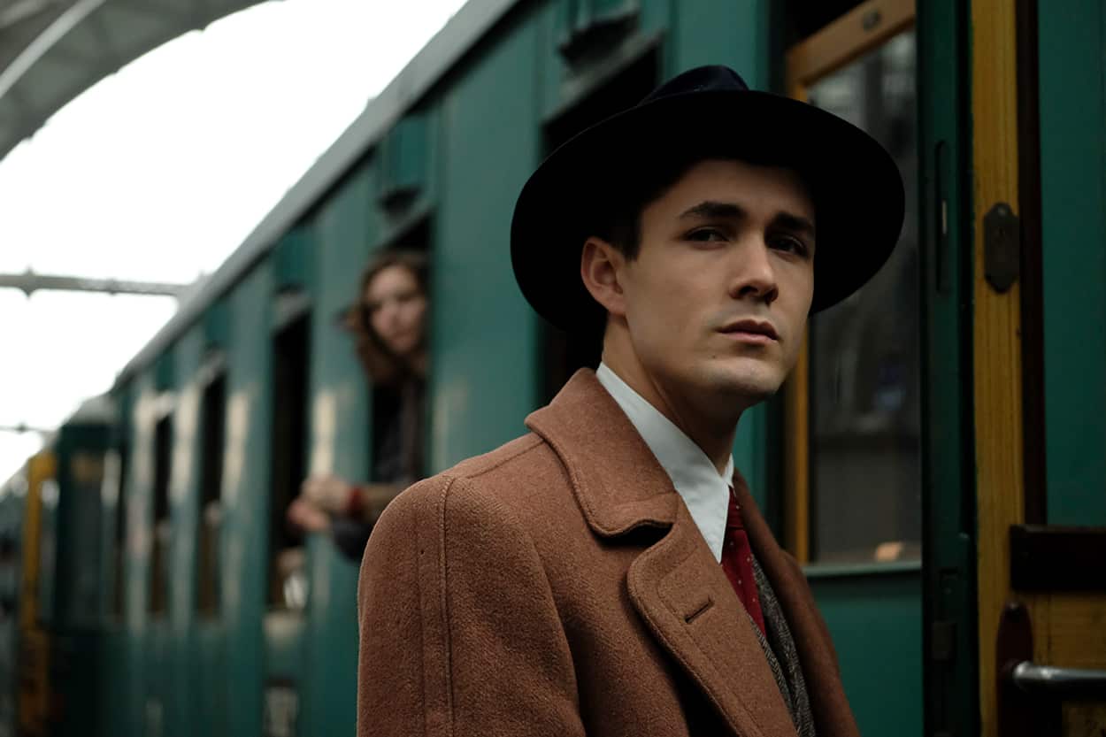 A man in a tan coat and a hat stands beside a train. 
