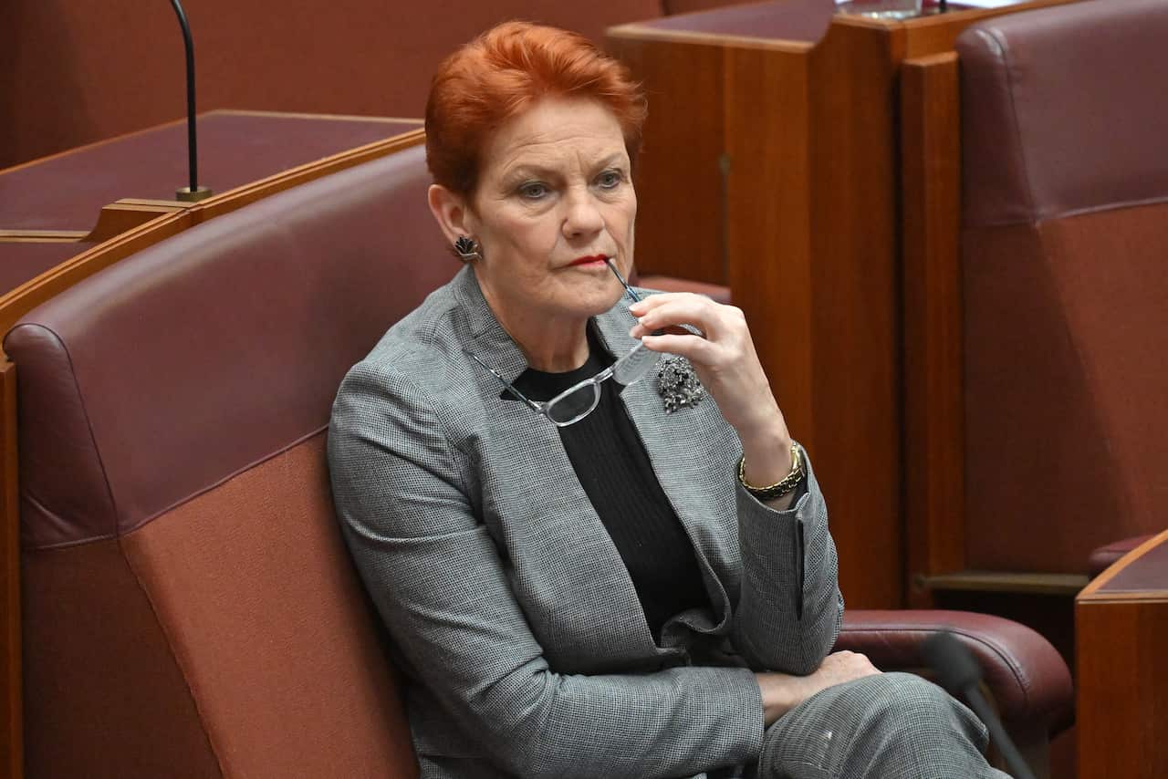 A woman in a grey suit is sitting on a red bench while chewing on the temple of her glasses.