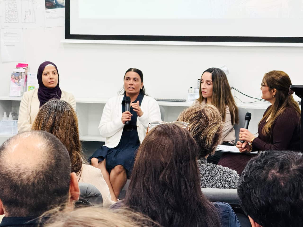 In the foreground are the backs of four people's heads. They watch four women holding microphones sitting in a row at the front of the room.