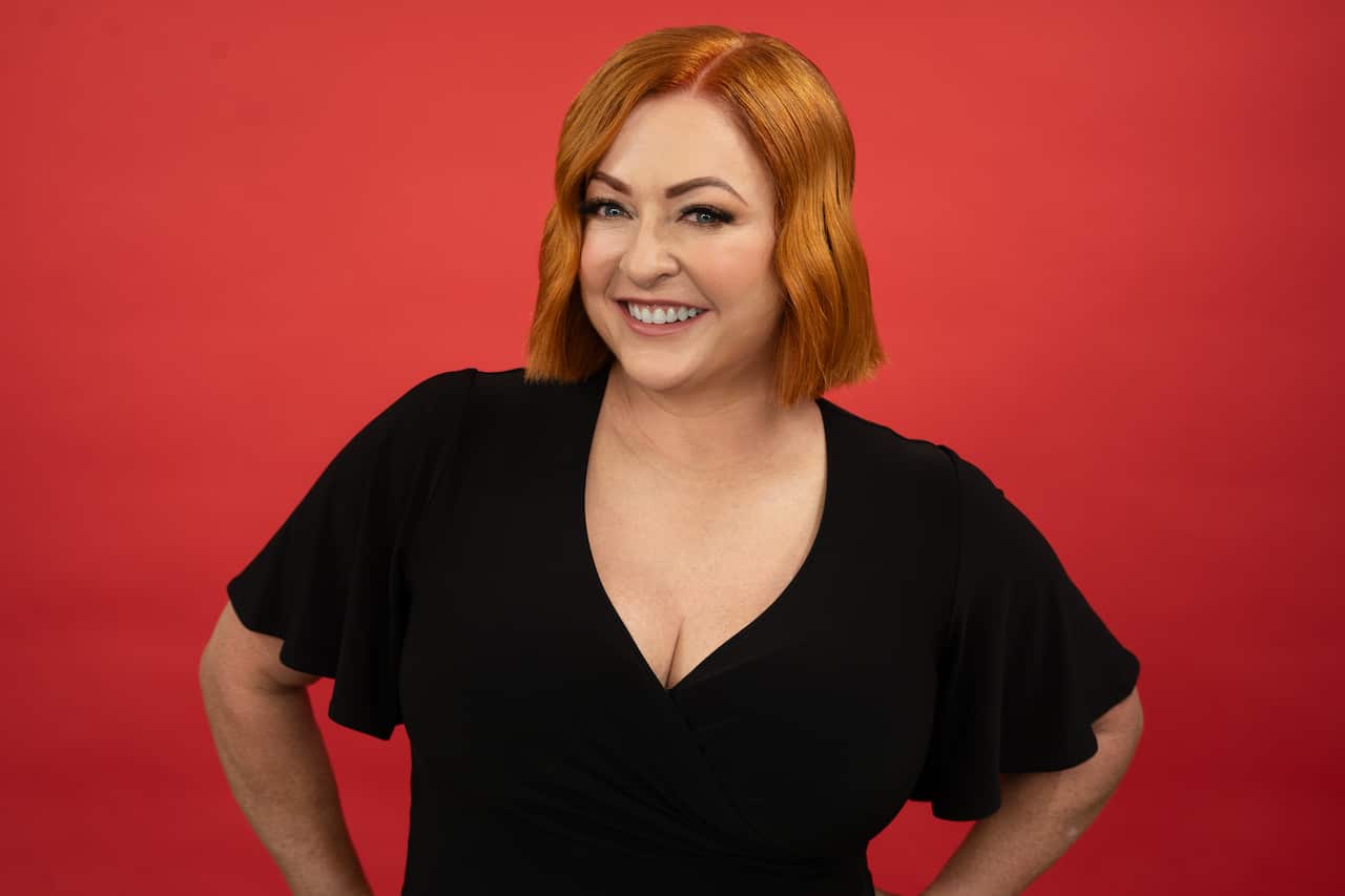 A woman in a black dress smiles at the camera against a red background.