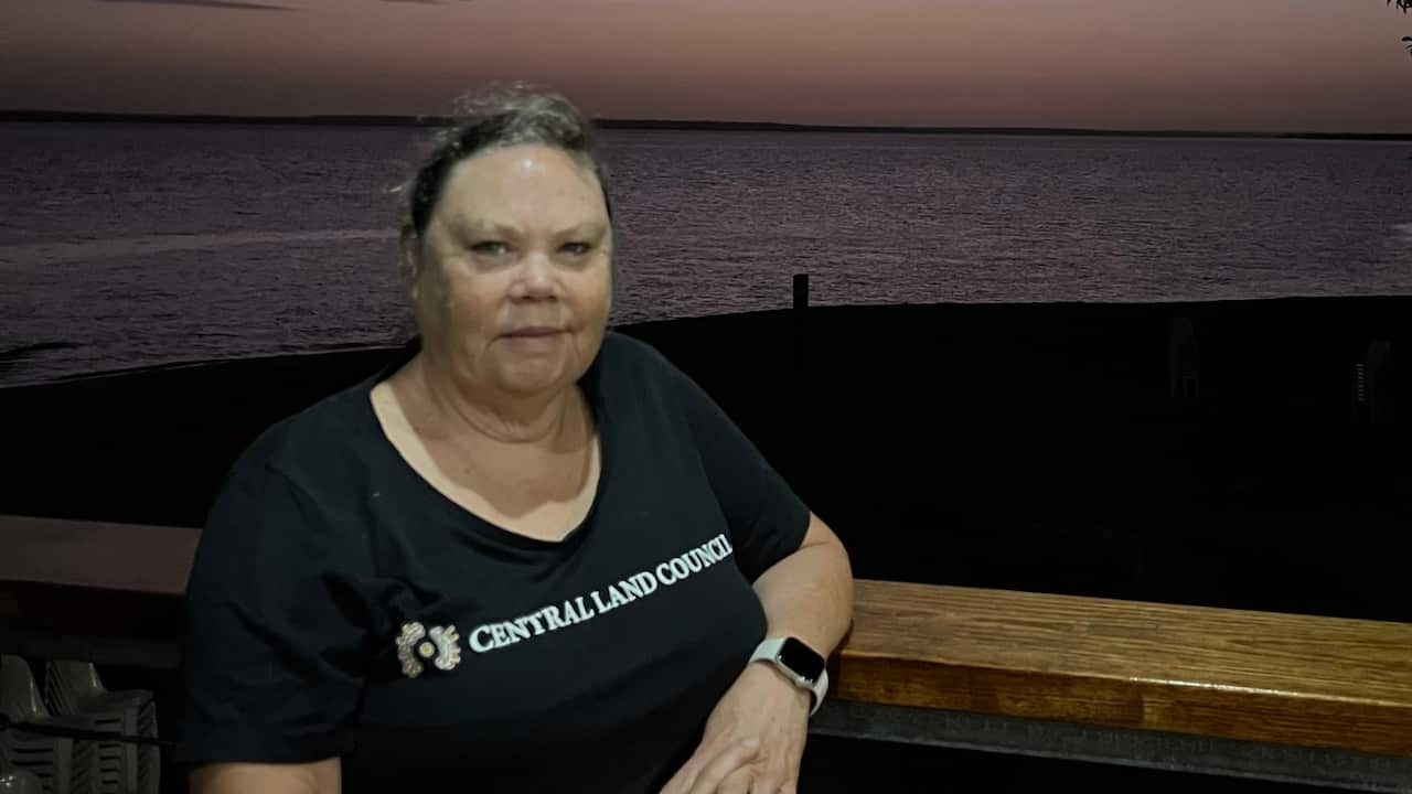 A middle aged woman standing in front of a beach at sunset time. She wears a black shirt that says Central Land Council