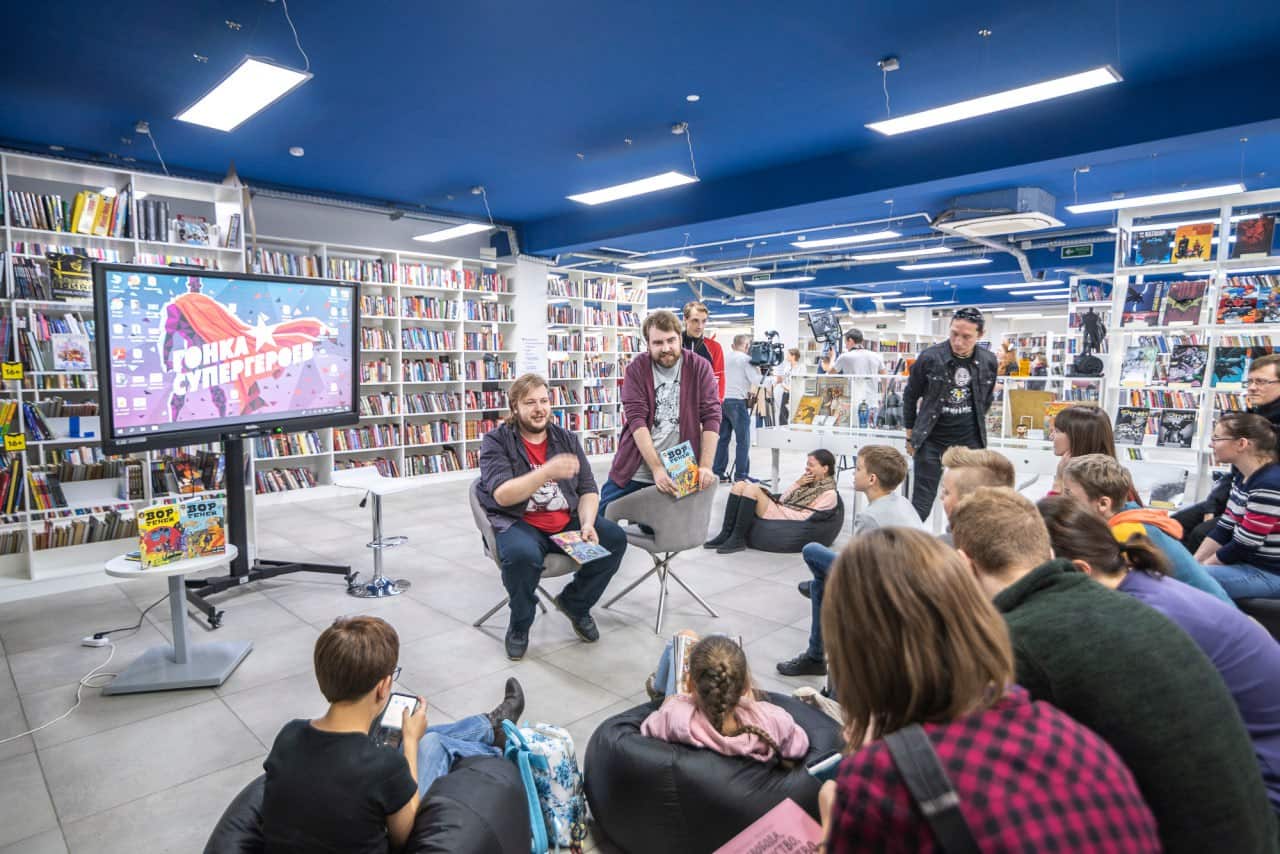 A group of people of various ages are seated on bean bags in a library, while two men on chairs address the audience. The walls are covered with bookshelves, and a TV screen behind the speakers displays the words 'Superhero race' written in the Russian language.