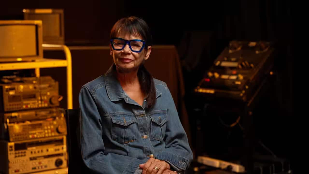 A woman wearing a denim shirt and blue glasses is sitting down, surrounded by old sound systems and televisions.