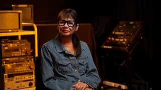 A woman wearing a denim shirt and blue glasses is sitting down, surrounded by old sound systems and televisions.