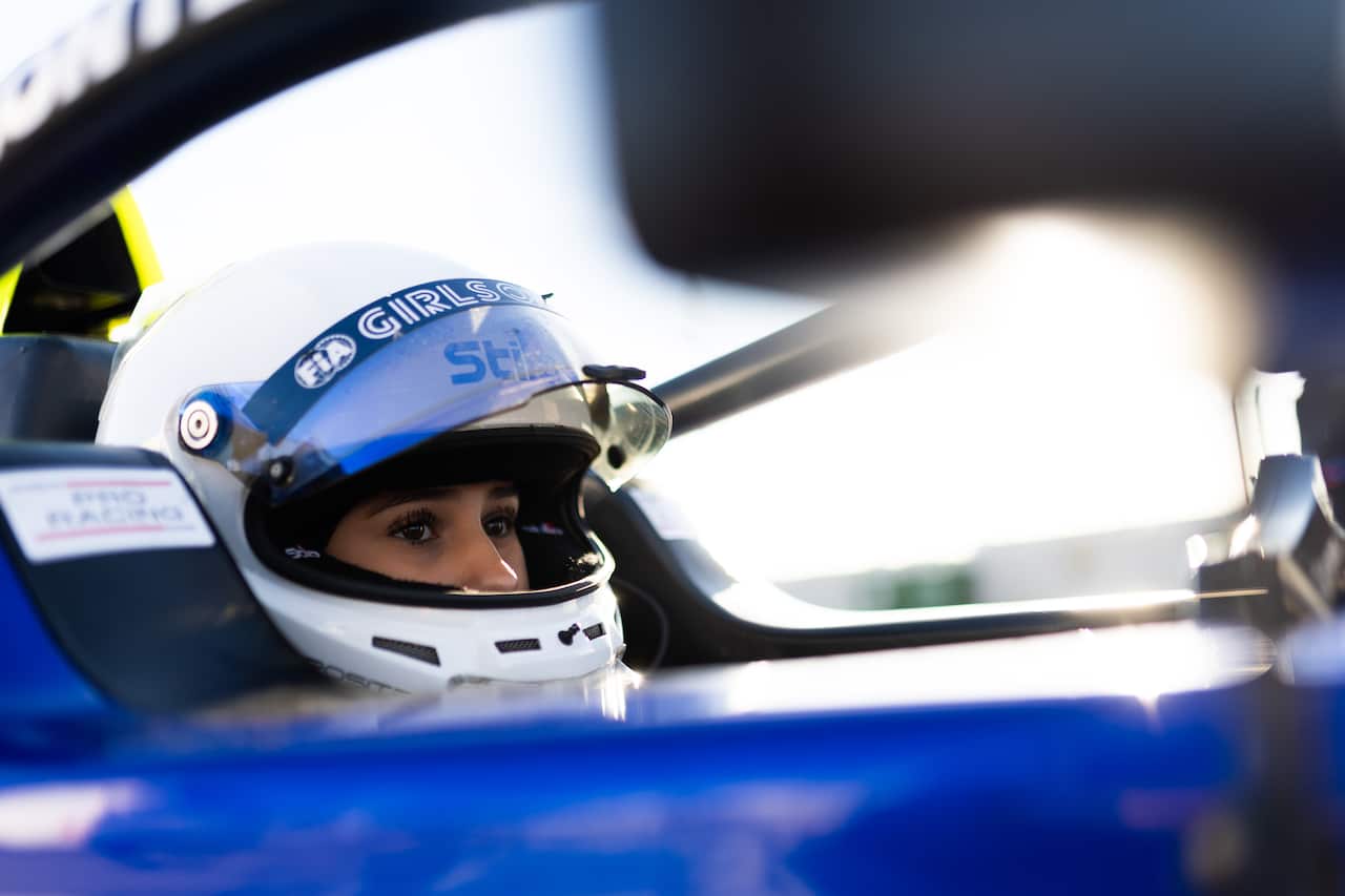 A girl wearing a white helmet looks out of the visor while sitting in a race car. 