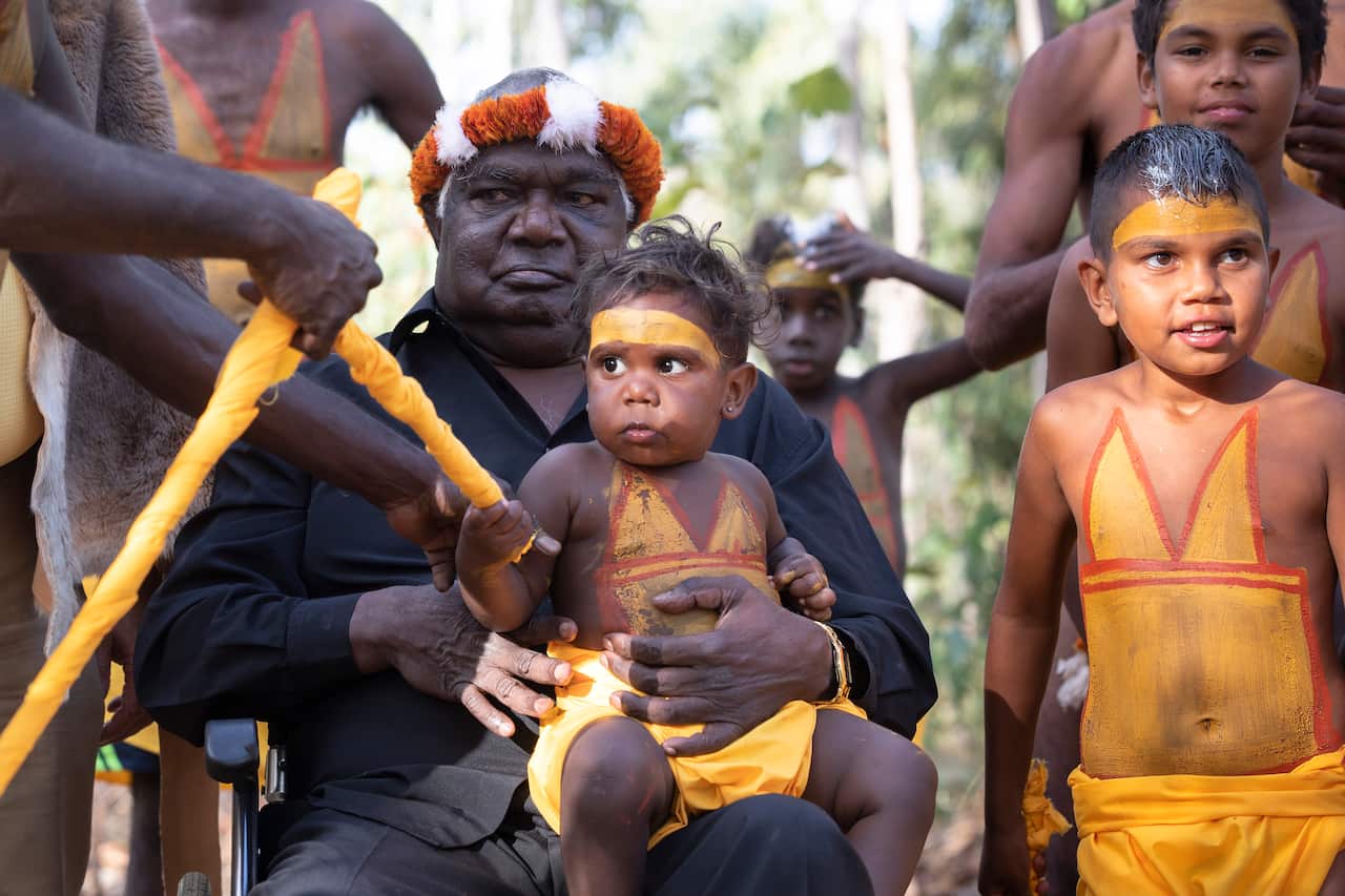 An Indigenous man wearing a black shirt and wreath sits with a young boy on his lap.