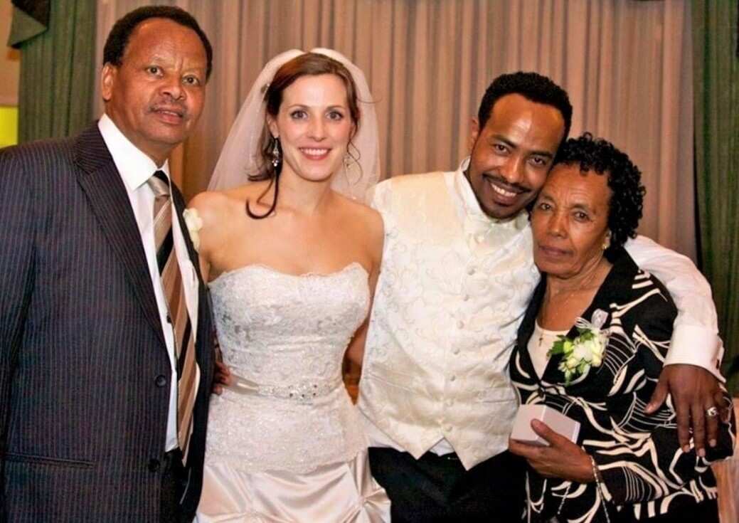 A man in a white satin vest hugs a woman standing next to a bride and an older man.
