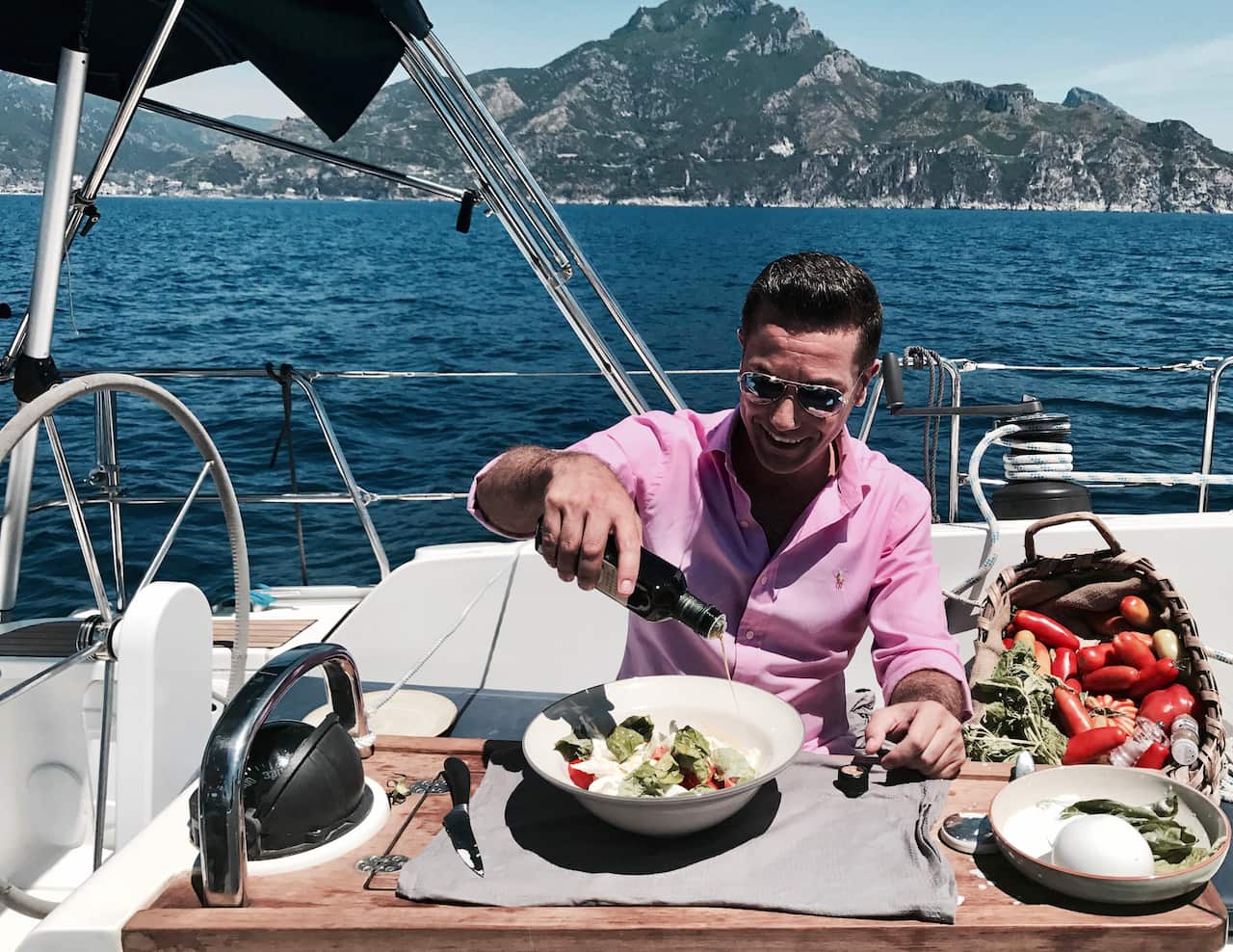 A man in a pink shirt and sunglasses sits at a table on a boat, making a salad. 