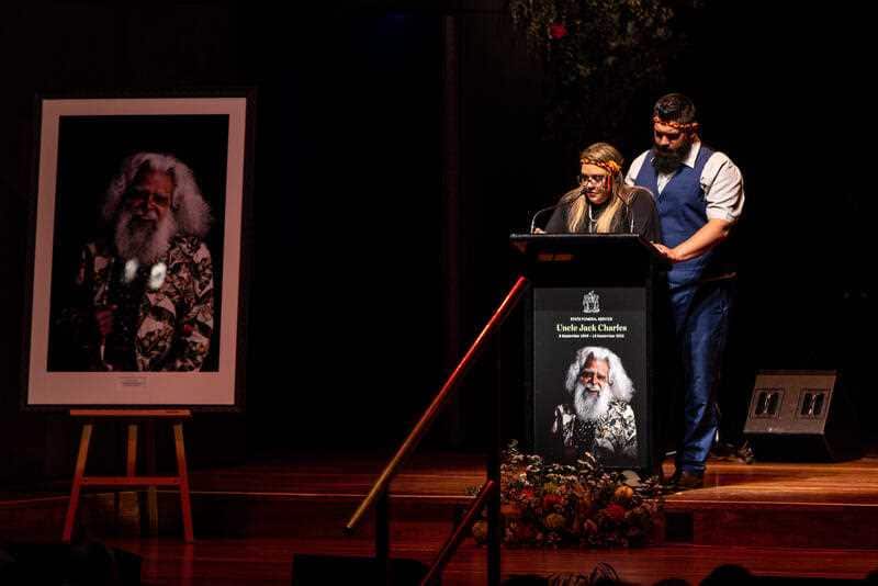 A woman and man stand behind a lectern bearing Jack charles' smiling face, a floral wreath at its foot.