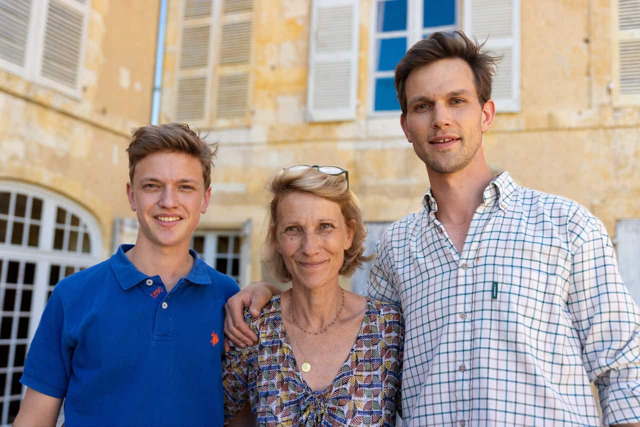 Two young men stand on either side of their mother. They are smiling or have neutral facial expressions, and have their arms around each other. Behind them is the wall of a manor house.  