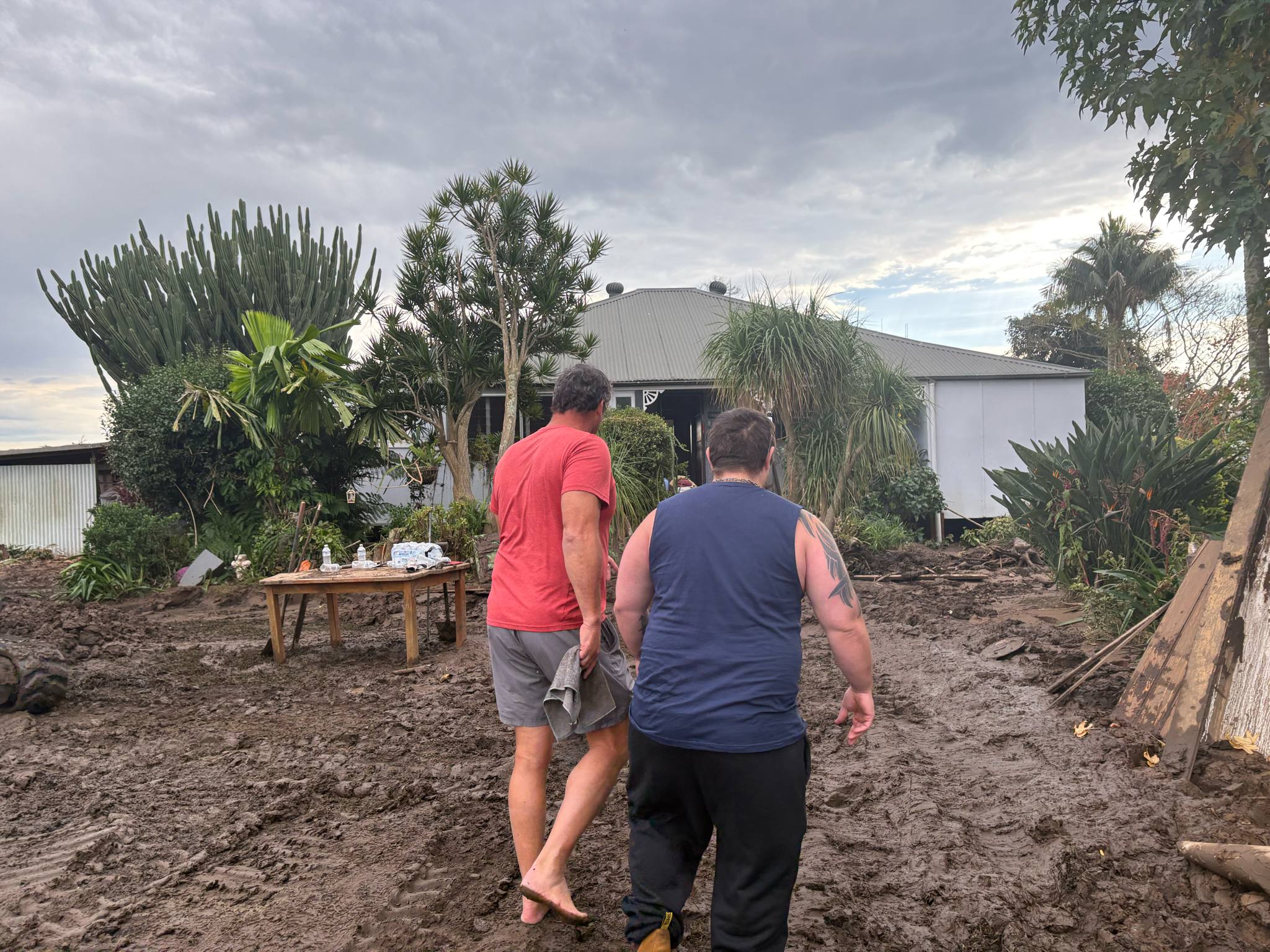 Two men walking up towards a house, their backs to the camera. The ground is covered in a thick muddy sludge. Debris is scattered everywhere. 