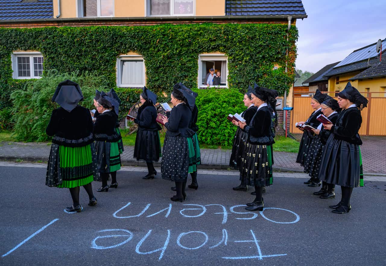 A group of women in black church costumes sing from hymn books in the street.