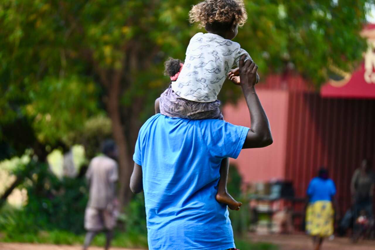 A Ramingining mother with her child on her shoulders walks to the supermarket