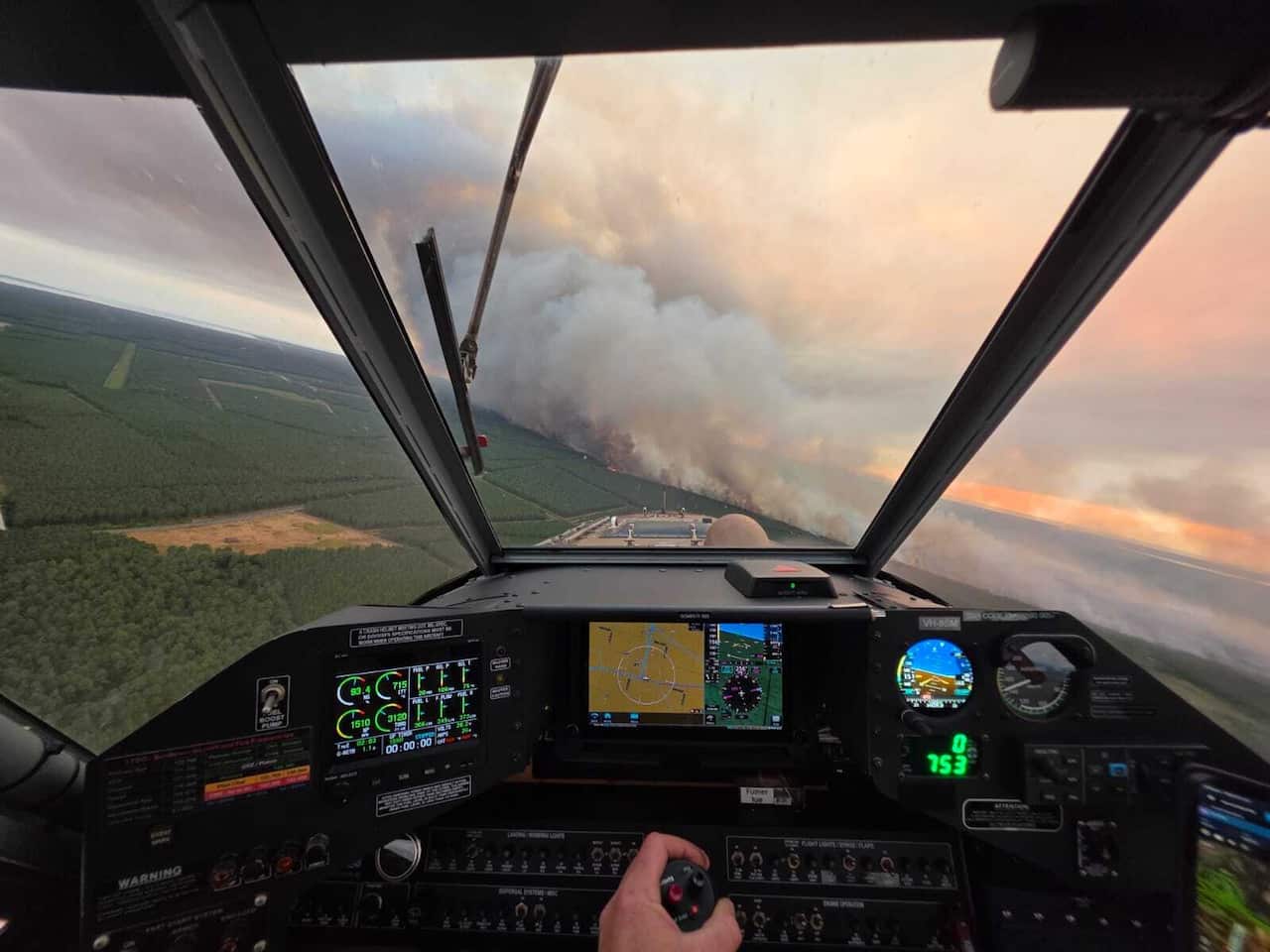 A pilot's perspective of flying a plane towards a wildfire burning in a forest. The cockpit is lit with screens and controls, and a hand controls the yoke