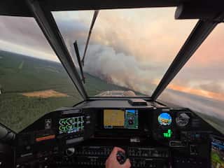 A pilot's perspective of flying a plane towards a wildfire burning in a forest. The cockpit is lit with screens and controls, and a hand controls the yoke