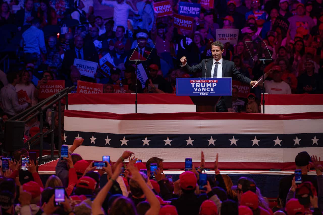 A man in a black suit addresses the crowd from a podium at a rally.