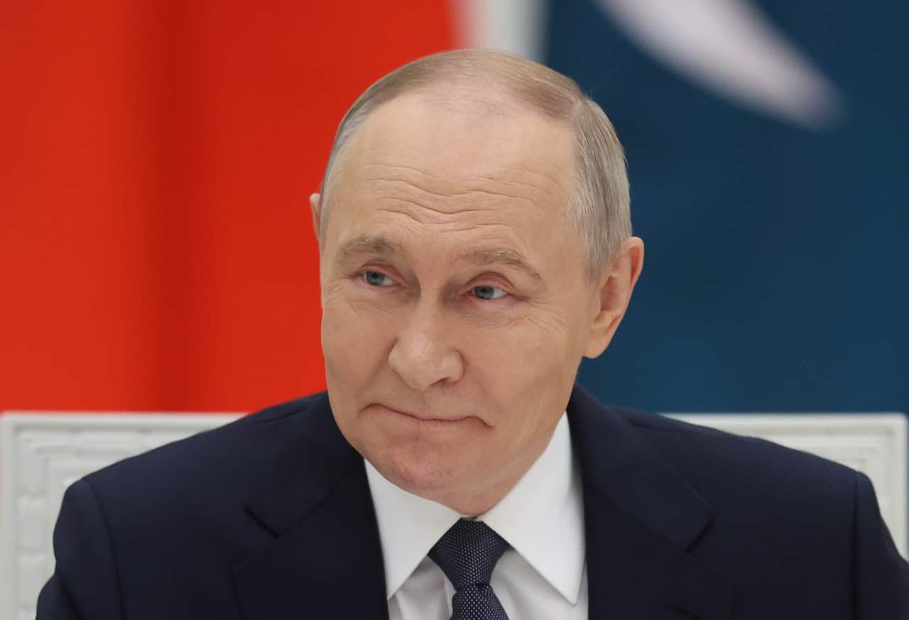 Vladimir Putin, wearing a black suit and white shirt, in front of a red and blue wall.