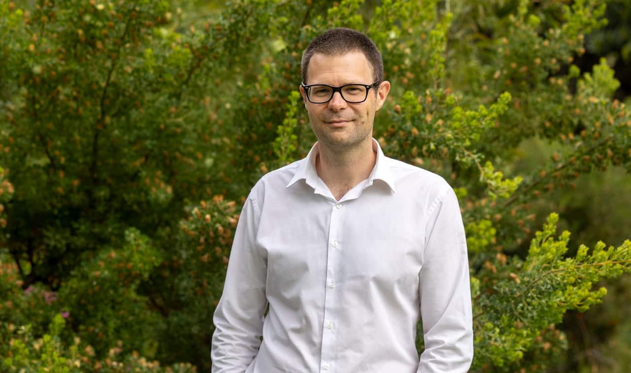 A man wearing a white shirt, standing in front of green bushes.