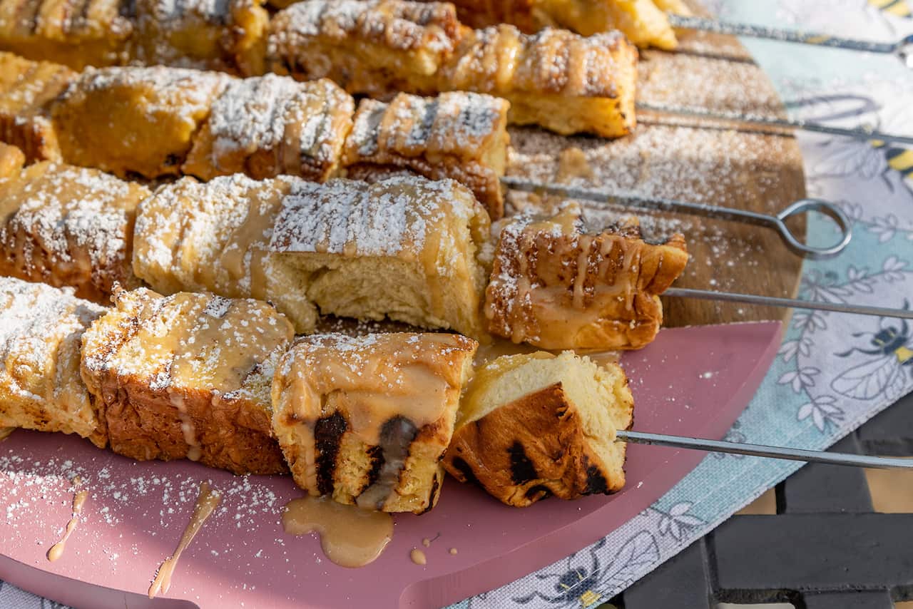 Five metal skewars, threaded with toasted brioche cubes, sit on a round wood and pink platter. The kebabs have been drizzled with a creamy-yellowish sauce and dusted with icing sugar. 