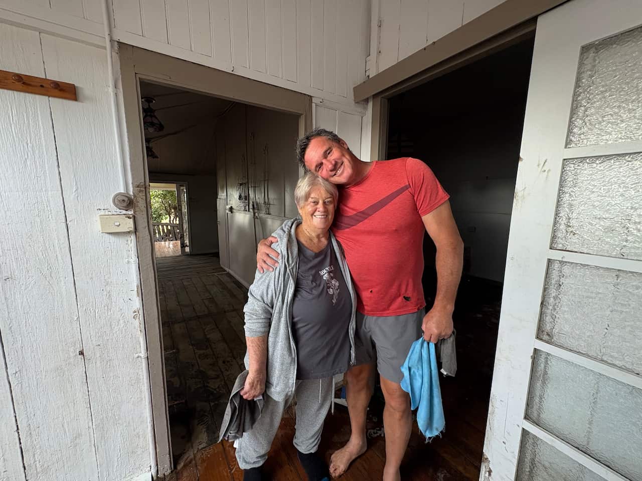 A very tall man wearing grey shorts and a pinkish red t-shirt stands on the right, holding a blue cleaning rag, with an older woman with short, white hair on the left dressed in all grey.