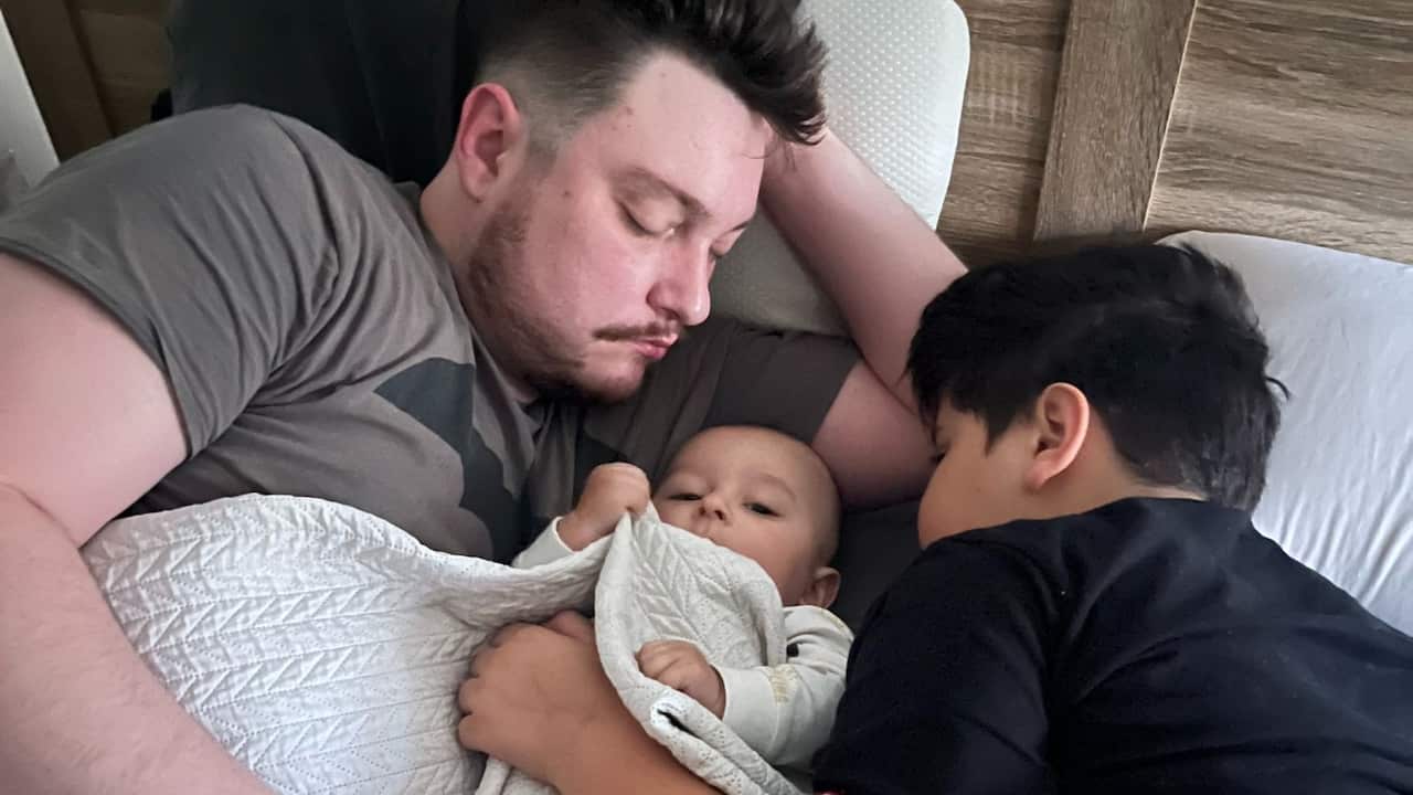 a dad lies asleep in bed with a baby and a young boy