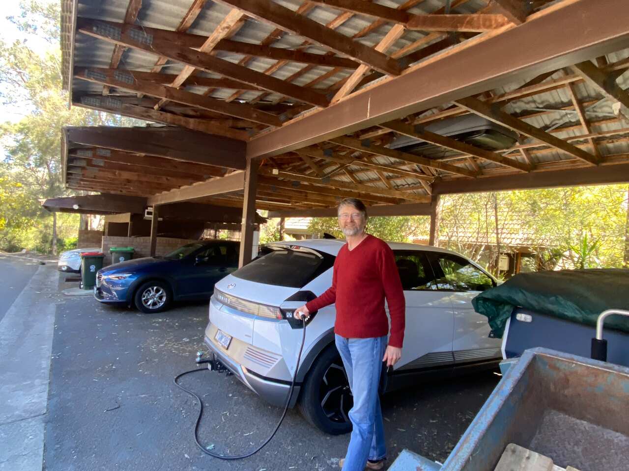A man holds a black cable that leads into his parked car.
