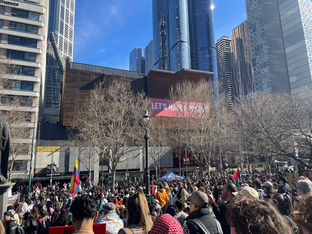 A crowd of protesters gather on a CBD street with skeletal winter trees in front of glimmering skyscrapers.