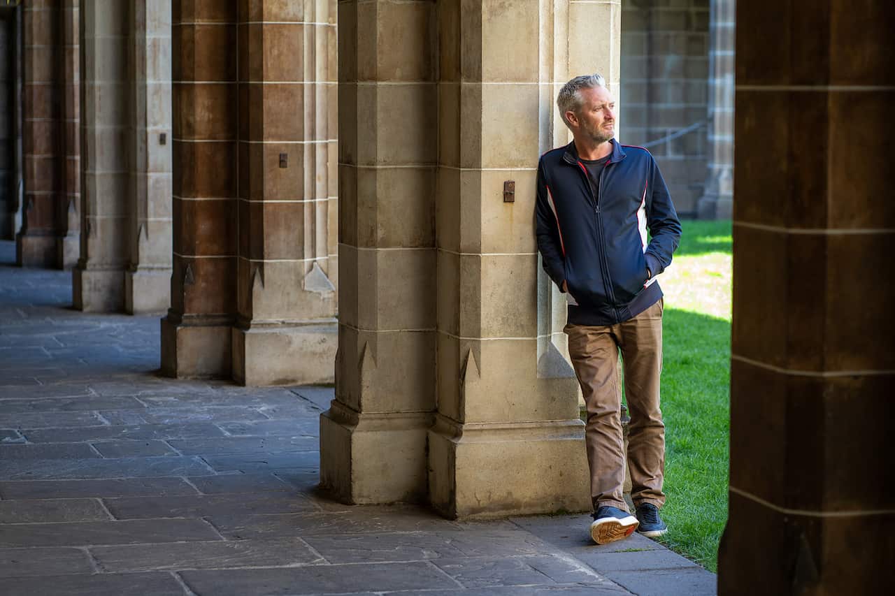 A man leans against a sandstone column, looking to one side. 