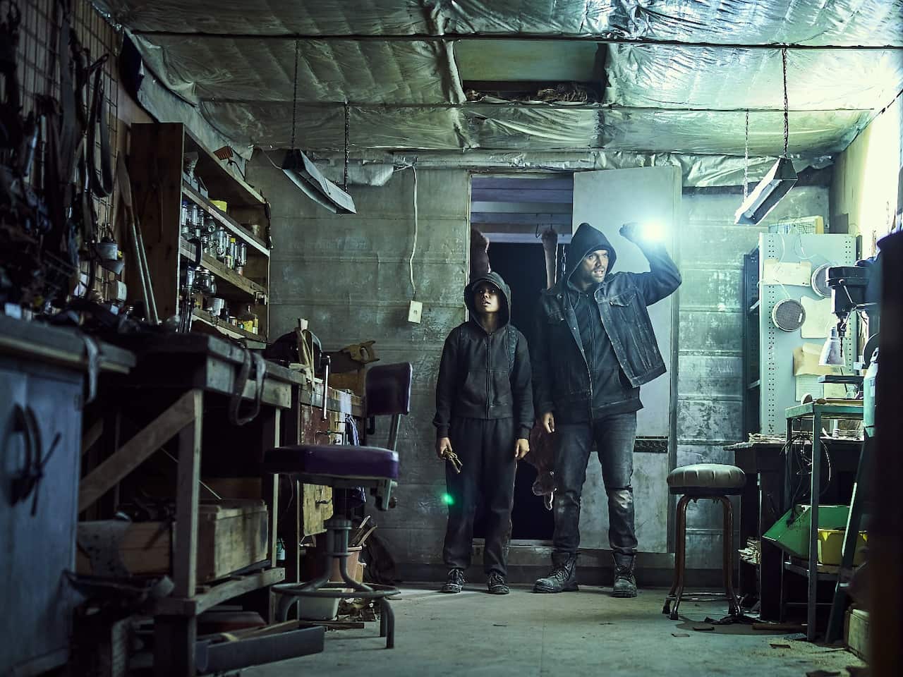 A young woman and a man stand in the doorway of a shed at night. The man holds up a torch to see inside.jpg