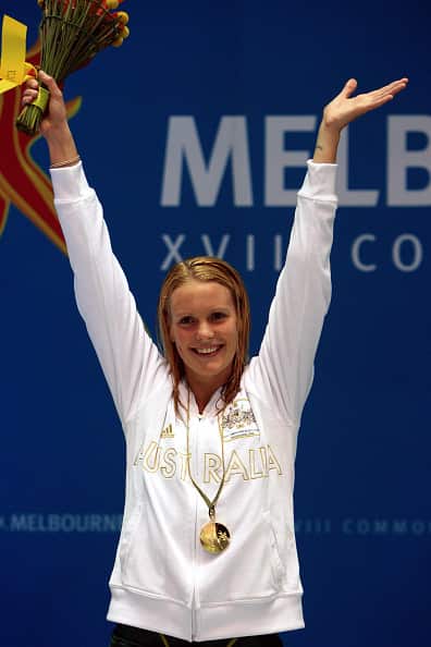 A smiling woman wearing a white tracksuit top and a gold medal around her neck stands with her arms raised.