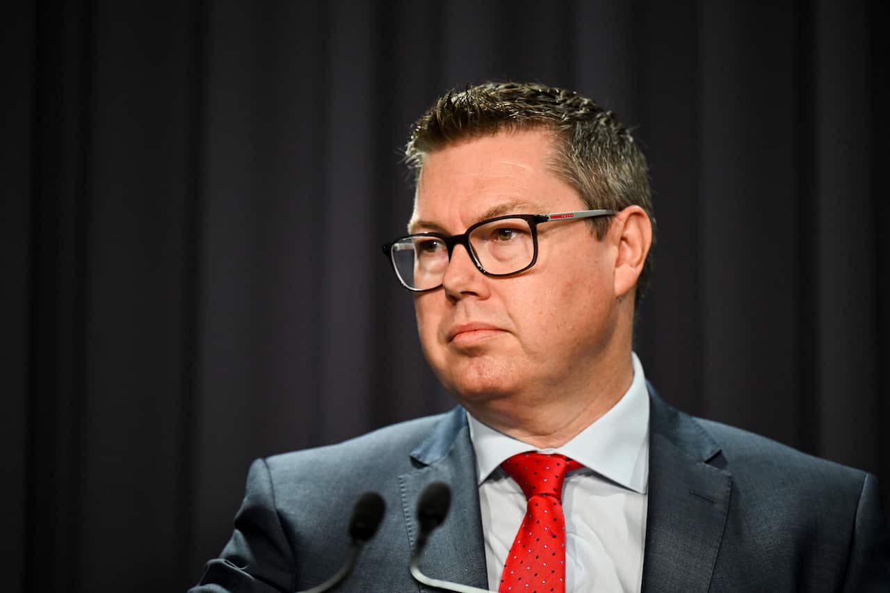 A man wearing black glasses and suit with a white shirt and tie standing in front of a black curtain