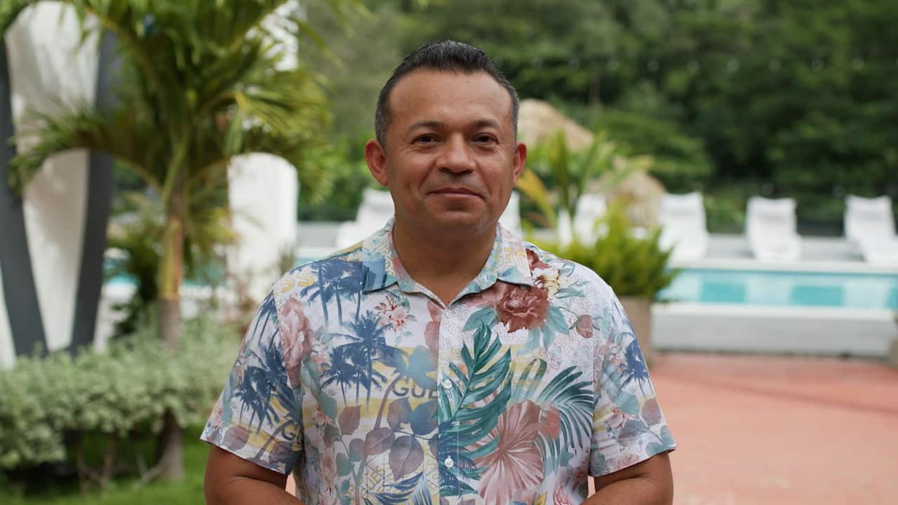 A middle-aged man wearing a short-sleeved tropical print shirt stands outside. There is a swimming pool in the background