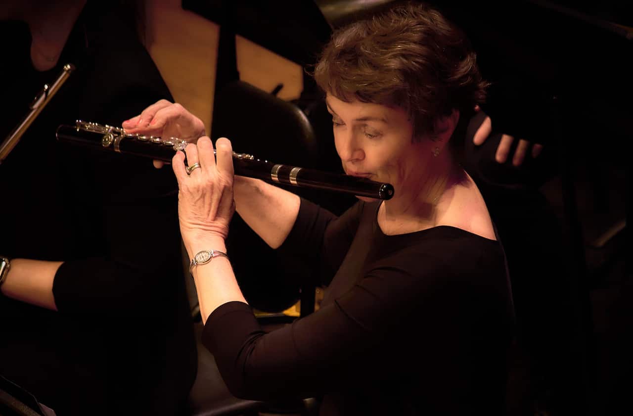 A woman with short hair and a black top playing the flute in an orchestra.