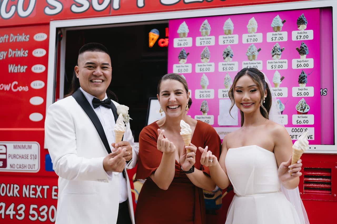 A bride and groom in wedding dress and suit, standing with another woman in a red dress, all holding ice creams in front of an ice-cream truck.