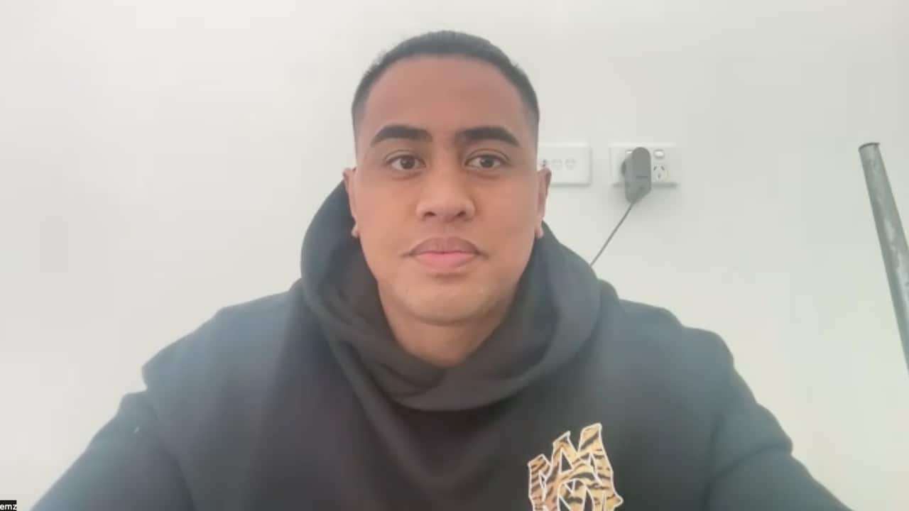A young man in a black hoodie sitting in front of a blank wall with a neutral facial expression.