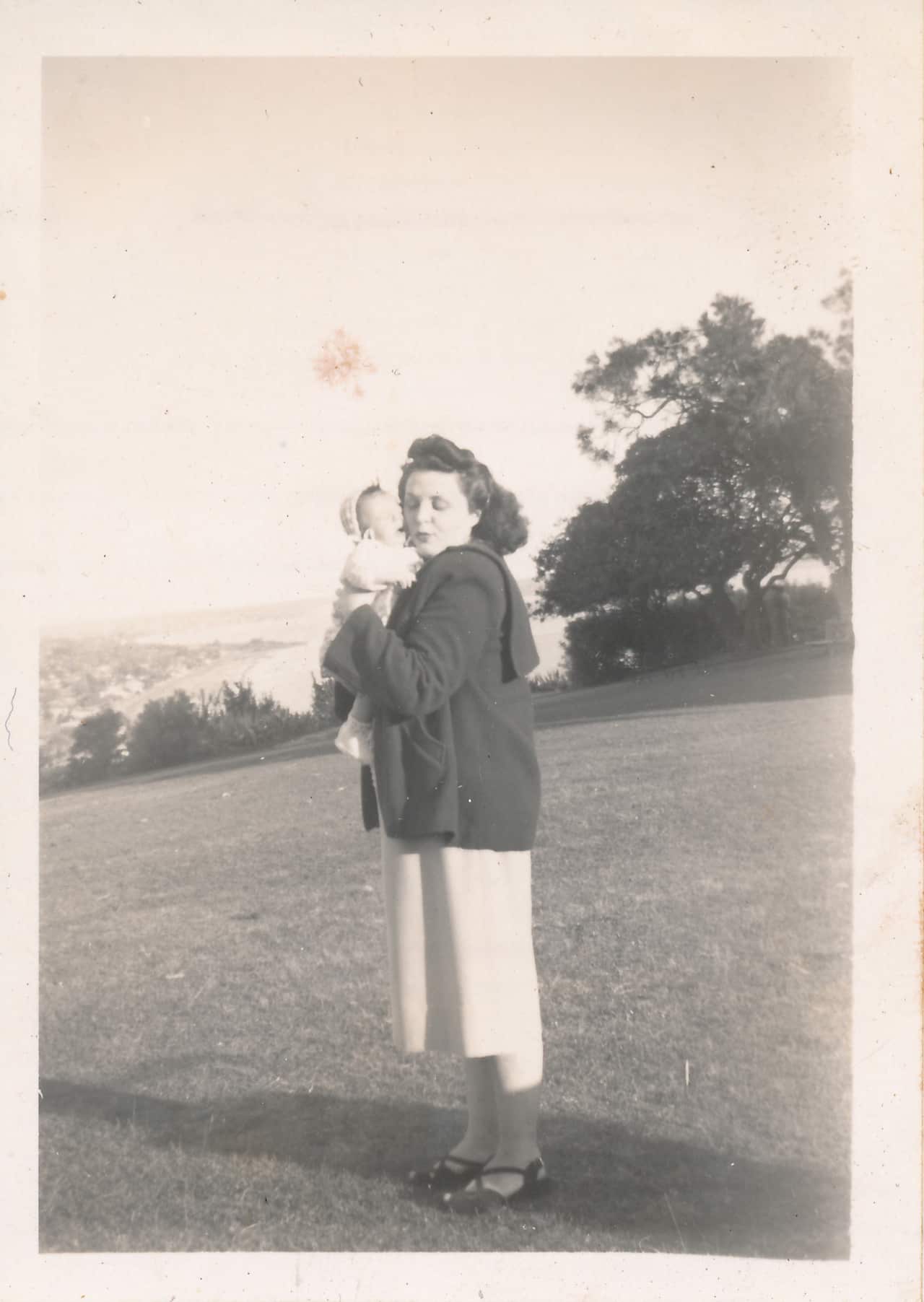 A black and white photo of a mother standing in a park holding her infant.