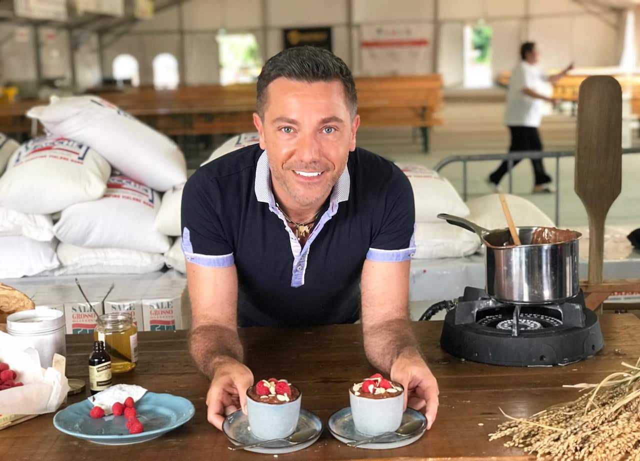 A smiling man leans on a table, with two desserts in small pots, on saucers, in his hands. Big bags of rice can be seen behind him. 