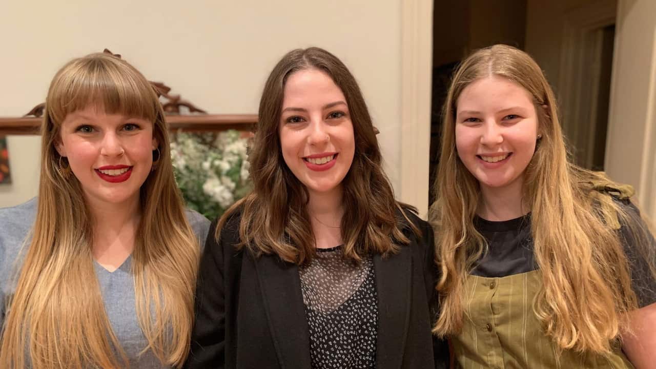 three young women smiling at the camera, the woman on the left has blonde hair and red lipstick and the woman on the middle wears a black top and has brown hair, the woman on the right has blonde hair
