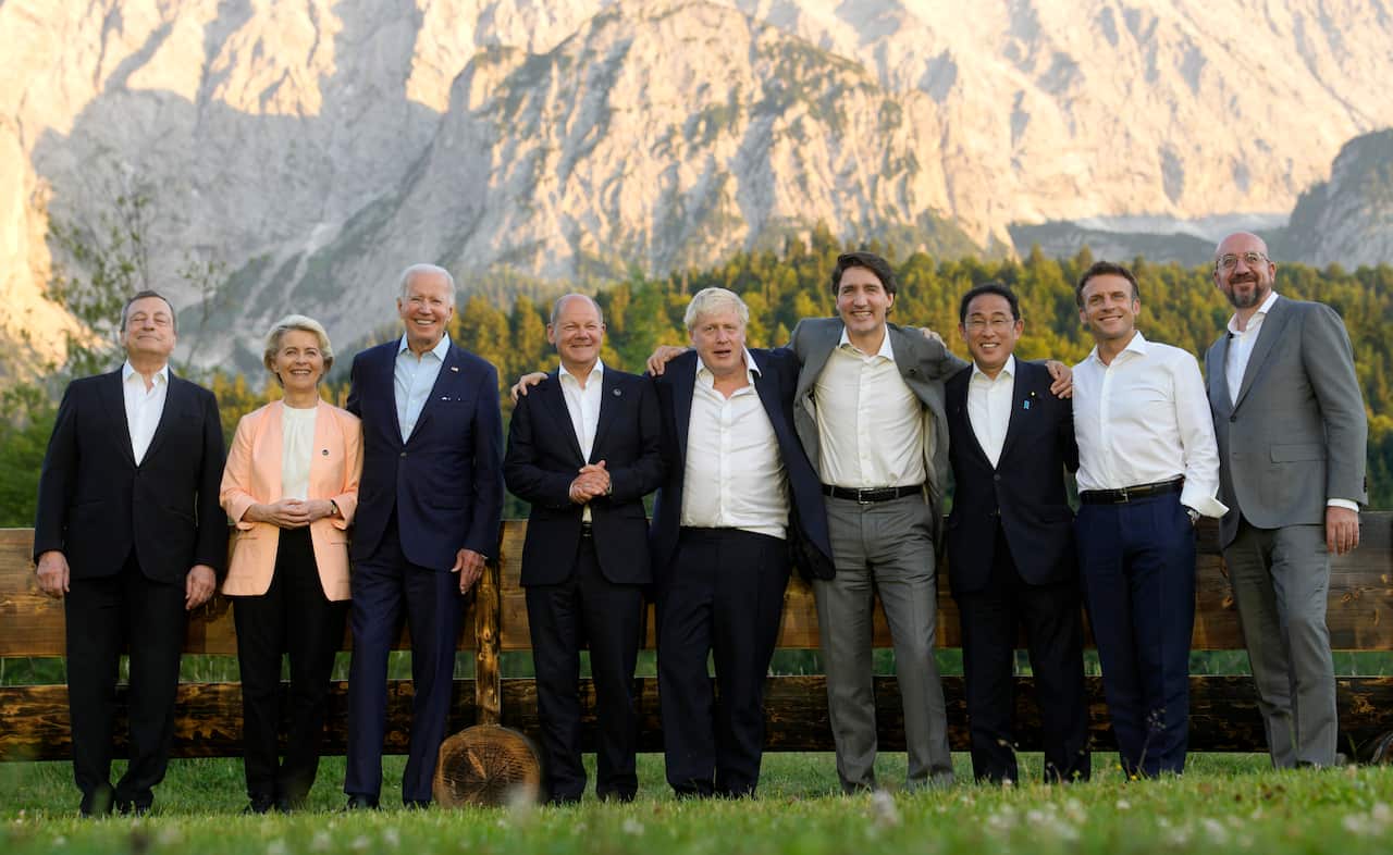 G7 leaders pose in a line together outside in a scenic area, in front of a mountain. 