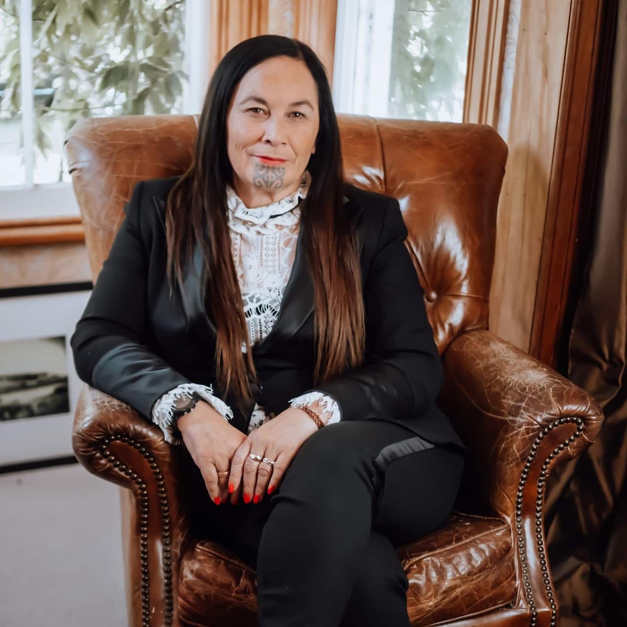 A middle-aged woman with long brown hair and a Maori Moko Kauae (facial tattoo) wearing a black suit sits on a brown leather chair. 