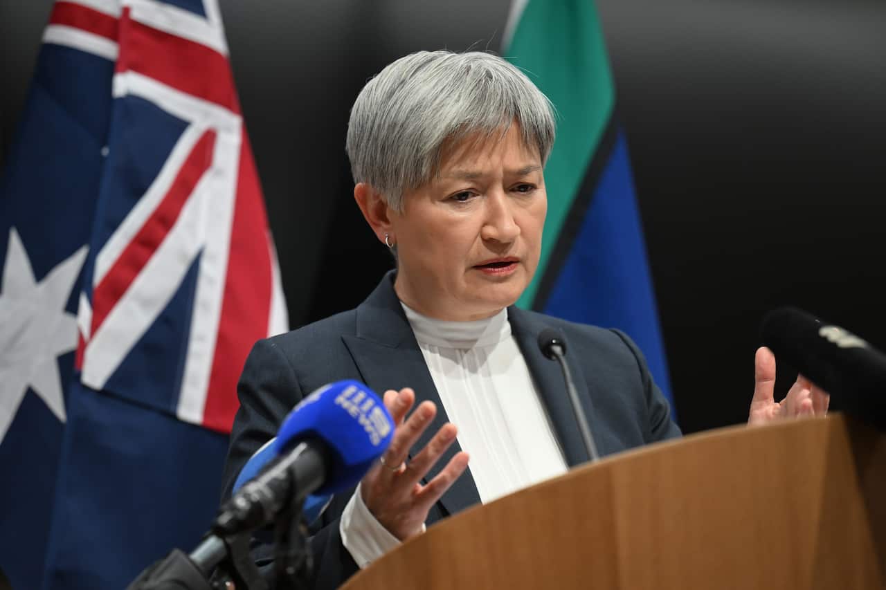 Penny Wong gesticulates as she speaks at a lectern, with Australian and Aboriginal flags in the background.