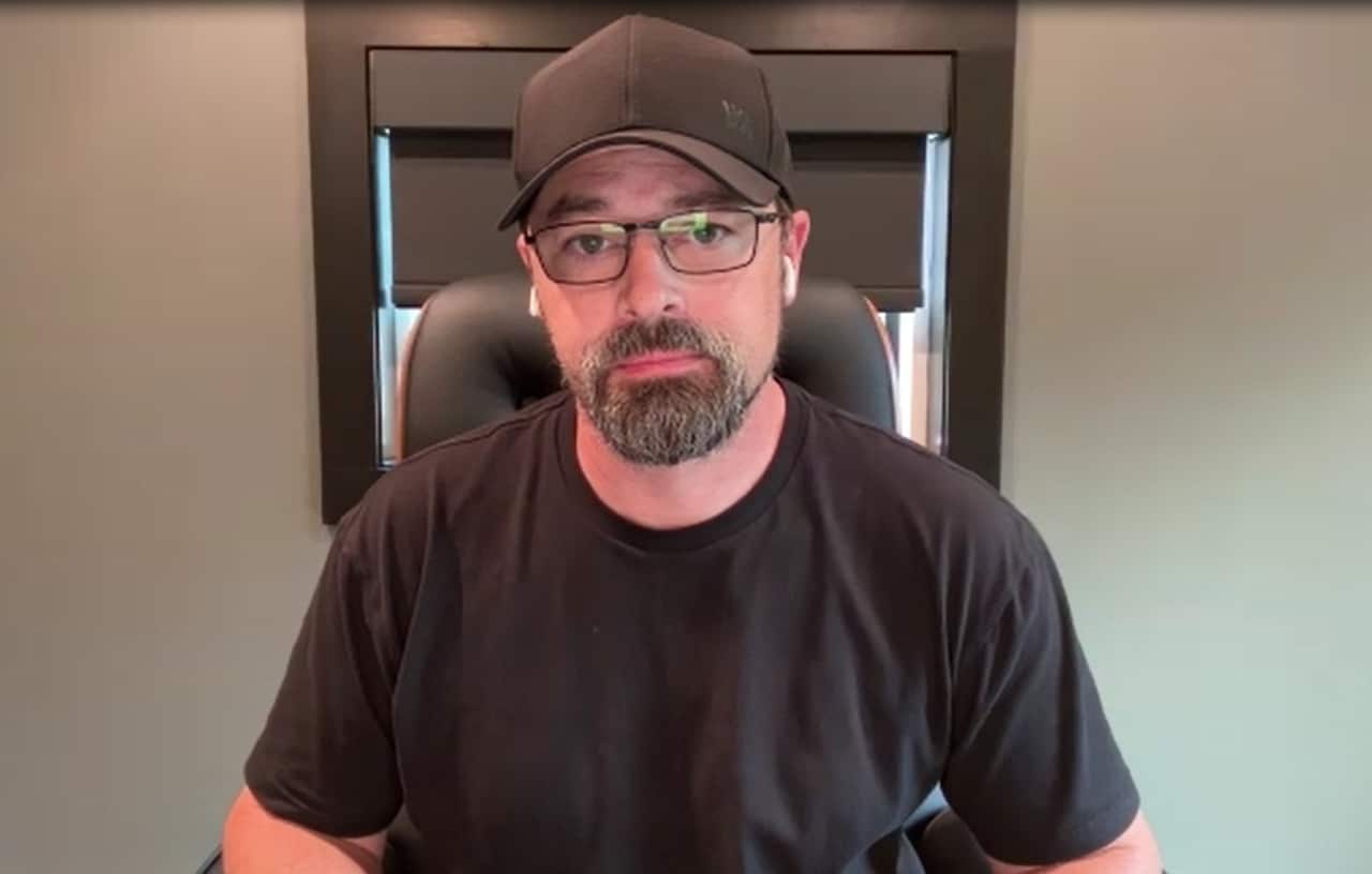 A middle-aged white man wearing a black cap, glasses, a black tee and a short beard. 