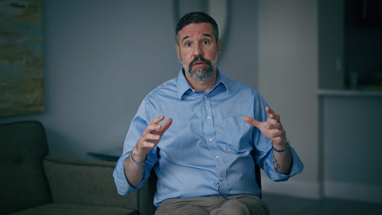 A medium shot of a middle-aged man with a salt-and-pepper beard wearing a blue button-down shirt, gesturing with his hands while speaking in an indoor setting.