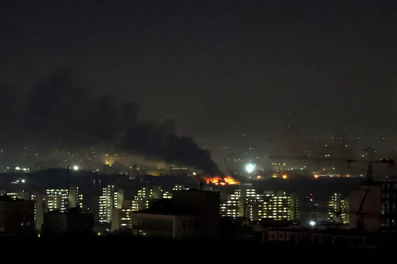 A nighttime cityscape shows thick black smoke and bright orange flames rising from a distant explosion site behind a row of illuminated residential buildings.