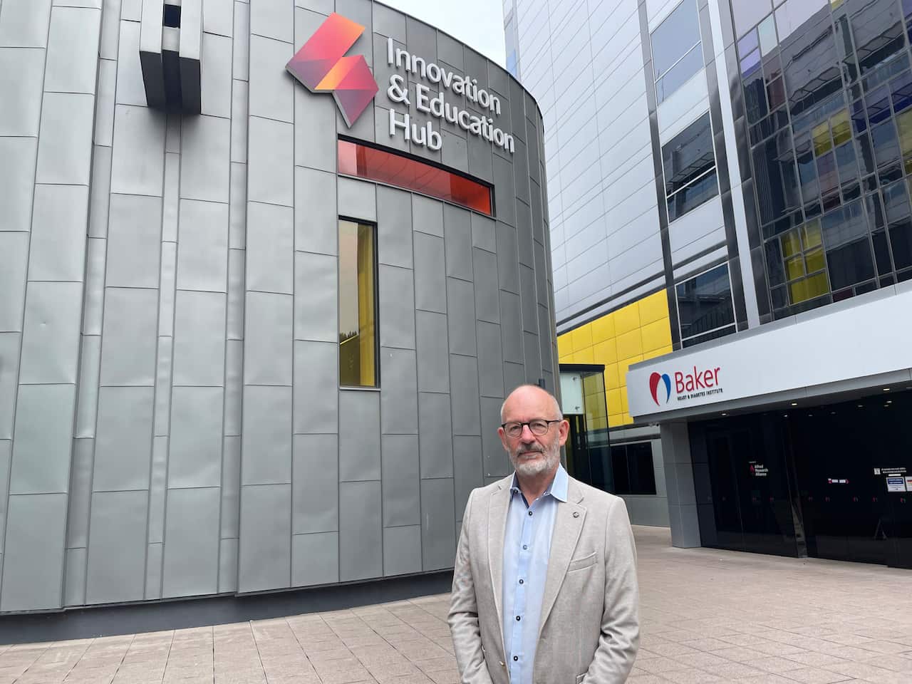 A man in a grey blazer and blue shirt standing in front of a building that reads "Innovation and Education Hub".