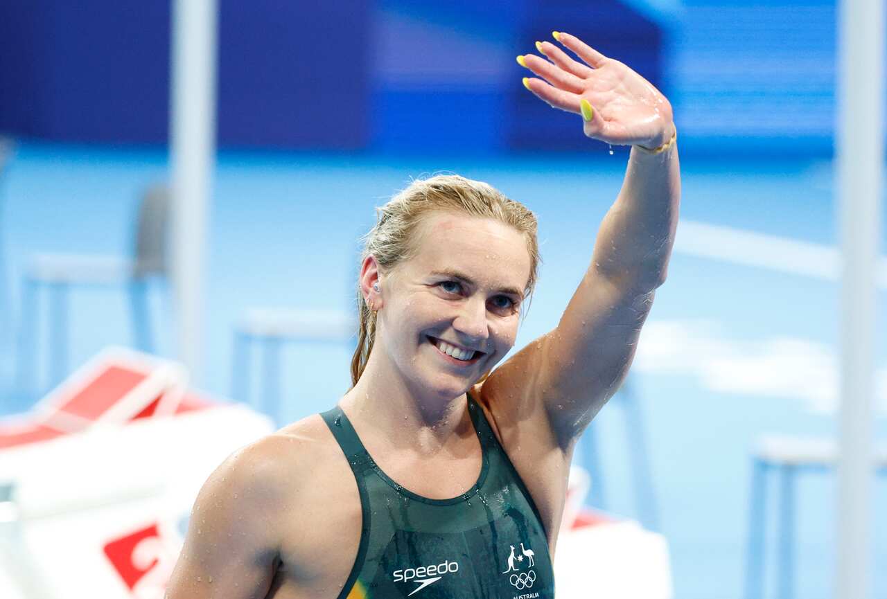A woman in a green swimming costume smiles and waves.