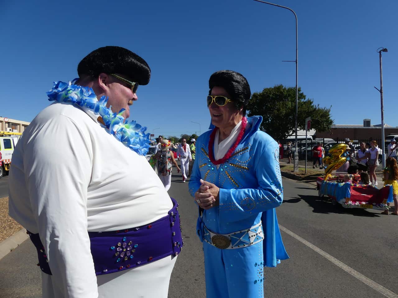Two men dressed like Elvis talking outside.