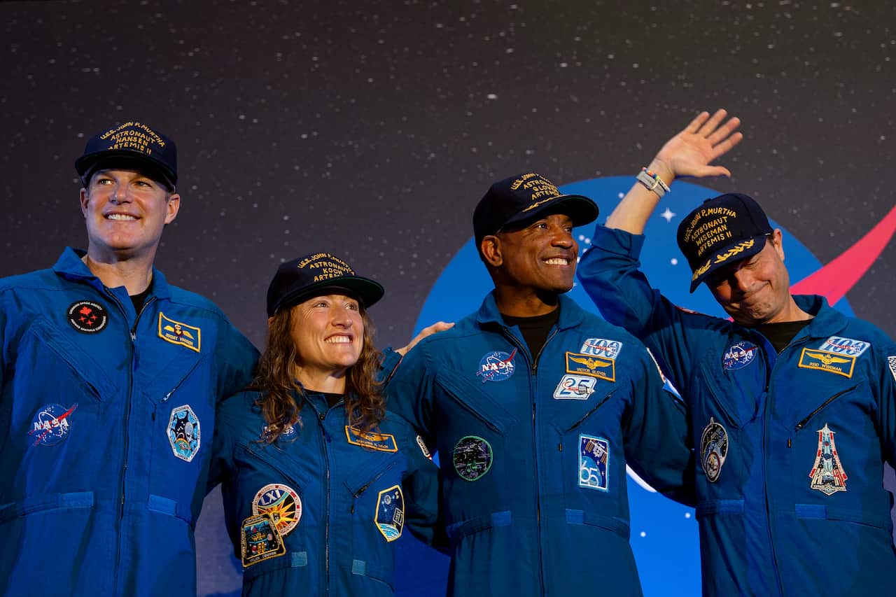 Two white men, a black man, and a white woman smiling and waving with their arms around each others backs. 
