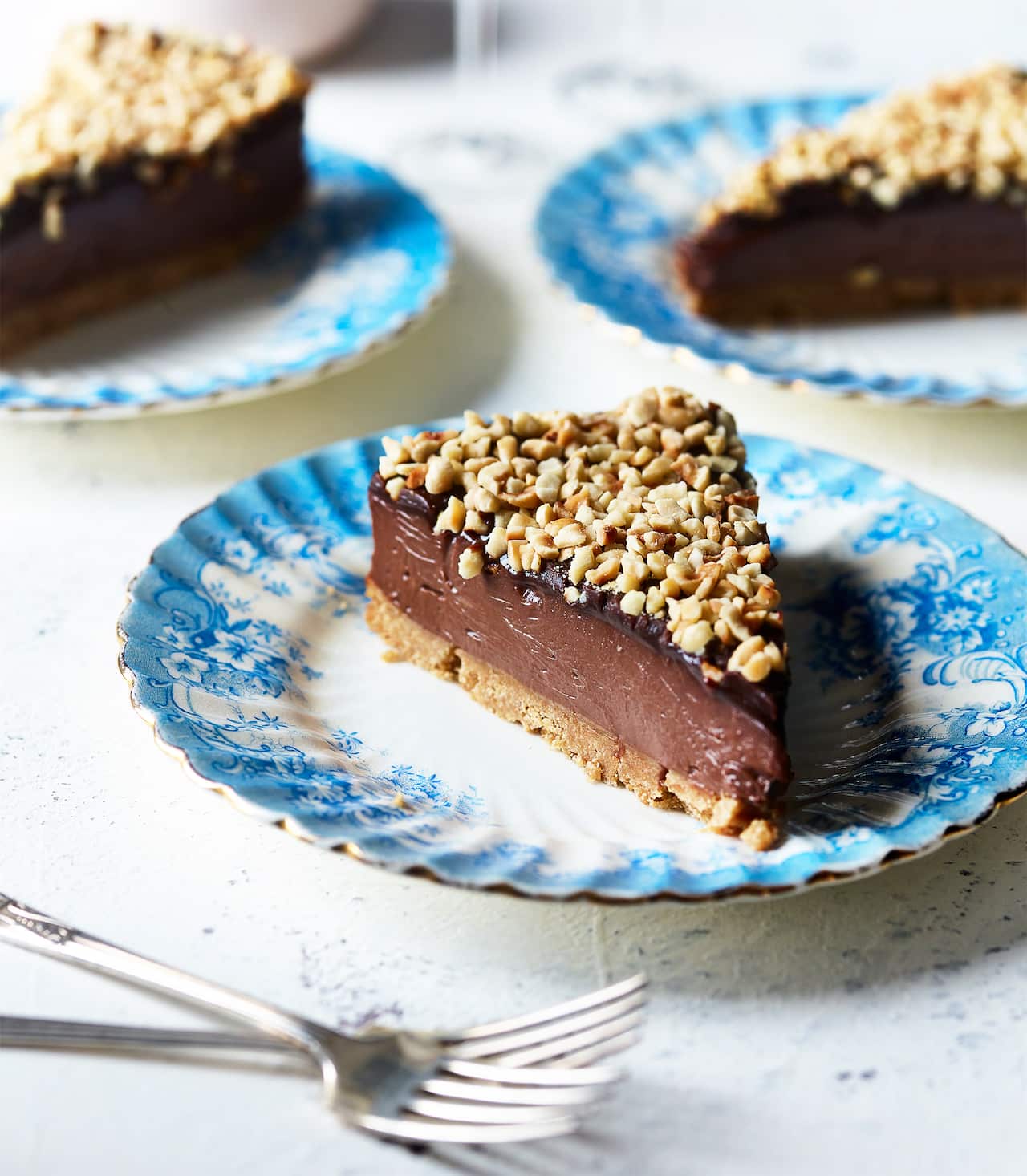 A wedge-shaped slice of cheesecake sits on a pretty plate with a textured and blue-patterned edge. The cheesecake is rich and lush, with a biscuit base, a deep middle layer and a thin darker top layer, sprinkled with chopped nuts. Two silver forks sit in front of the plate. Two more plates, with slices of cheesecakes, can be seen in the background.